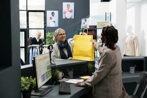 Elderly shopper buying fashionable clothes in boutique shop, taking shopping bag from store employee. Old woman standing at cash register paying for stylish clothes at pos terminal photo