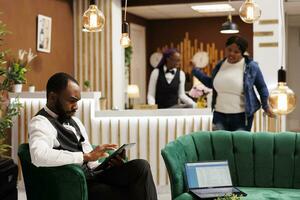 African American male hotel manager wearing uniform sitting in lobby using digital tablet managing bookings, bellboy holding electronic device entering details in left luggage register photo