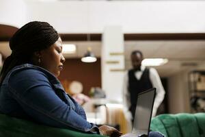 Serious focused African American woman traveler looking at laptop screen while sitting at hotel lobby, female guest using computer for online checkin. Remote work and traveling concept photo