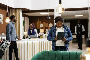 African American woman tourist standing in hotel lobby with digital tablet in hands doing selfcheckin. Female traveler completing registration procedure with electronic device, booking room online photo