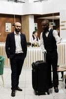 Bellboy leading businessman to room, carrying luggage for him and ensuring pleasant stay at hotel. Bellhop offering luxury concierge services to guest travelling for work, front desk. photo