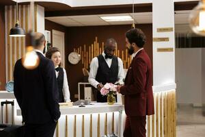 Professionals greeted by hotel staff at front desk, businessmen in suits arriving at luxury resort. People travelling for work partnership, doing check in procedure at reception. photo