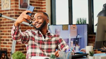 Joyful cheerful african american man taking selfie while smiling and showing thumbs up sign, buttoning up shirt. Person in modern house capturing photo using smartphone with background tv noise