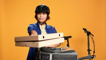 Courier woman isolated over orange studio background bringing pizza to customer. BIPOC bike rider delivering order to client, taking fast food boxes out of thermal backpack photo