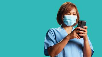 Cheerful healthcare expert texting friends online while taking clinic job shift break, close up. Happy nurse typing message on phone, wearing antibacterial face mask, isolated over studio background photo