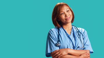 Joyful experienced asian nurse folding arms, smiling while looking at camera. Portrait of happy healthcare expert wearing protective scrubs, isolated over blue studio background photo