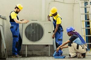 Seasoned mechanic coworkers installing outside air conditioner for customer. Adept repairmen commissioned to optimize new HVAC system's performance, ensuring it operates at maximum efficiency photo