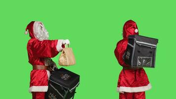 Festive man in santa claus suit deliver food with paper bag, posing with thermal backpack over greenscreen backdrop. Person in traditional holiday costume spreading christmas spirit. photo