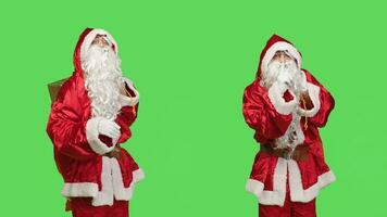 Father christmas shows mute symbol, espressing privacy and silence over greenscreen in studio. Santa claus with gifts bag doing hush sign to keep secret, private seasonal winter character. photo