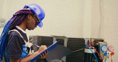 Skilled african american expert commissioned for outdoor air conditioner annual check up, writing findings on clipboard. Seasoned wireman doing hvac system investigation of damaged parts photo