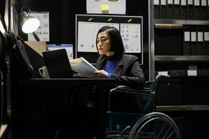 Asian accountant in wheelchair checking annual profit revenue documents before business consulting meeting. Woman with paraplegia in bureaucratic workplace filled with flowcharts and folders photo