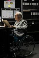 Senior businessman executive in wheelchair checking accountancy budget plan data paperwork. Experienced bureaucratic employee in depository filled with document folders chart reports photo