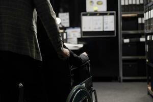 Paralyzed private detective wheelchair user and assisting coworker in criminal cases archive room. Investigator with paraplegia surrounded by criminology folders on cabinet shelves photo