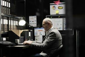 Focused elderly professional accountant checking administrative analytical data in file room surrounded by accountancy statistic charts. Businessman in bureaucratic office filled with invoice folders photo