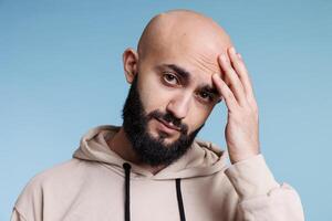 árabe hombre conmovedor cabeza en dolor mientras teniendo migraña y mirando a cámara. joven persona sufrimiento desde dolor de cabeza mientras participación mano en frente y posando para estudio retrato foto