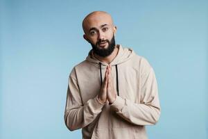 Hopeful arab man looking at camera with folded hands in pleading gesture. Young handsome person wearing casual hoodie praying, making praise sign with arms studio portrait photo