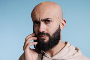 confuso árabe hombre respondiendo teléfono inteligente llamada y ceñudo Cejas retrato. joven calvo barbado persona hablando en móvil teléfono y mirando a cámara con incierto expresión foto