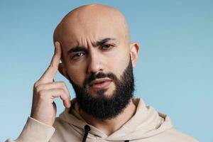 Arab man rubbing temple while suffering from migraine attack portrait. Young adult arabian bald bearded person grimacing while experiencing headache and looking at camera photo