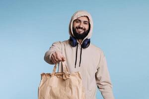 Cheerful smiling arab man in hood giving paper bag with take out food portrait. Restaurant delivery service courier holding package with takeaway lunch and looking at camera photo