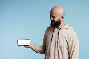 Arab man holding mobile phone and looking at empty white screen with copy space. Cheerful carefree person showing smartphone with blank touchscreen in horizontal mode for advertisement photo