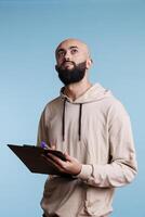 Pensive arab man holding clipboard and contemplating checklist while looking upwards with thoughtful facial expression. Young person thinking while taking notes and planning photo