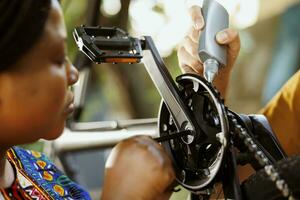 africano americano mujer examina bicicleta pedales para daños y perjuicios a reparar mientras caucásico masculino mano usos especializado lubricante en cadenas para ciclo eficiencia. dos joven activo individuos trabajando en moderno foto