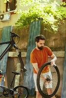 Energetic caucasian male dismantling damaged bicycle tire to repair and replace with new. Young athletic man grasping and carrying bike wheel for further maintenance in home yard. photo