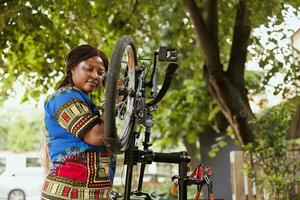 Focused healthy african american woman mending her own bicycle in yard and performing annual outdoor maintenance using expert tools. Female cyclist inspects and repairs bike pedals. photo