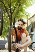 Active and committed man crouching to repair bike wheel rim in home yard using professional tool. Healthy male cyclist working on dismantled bicycle tire outside using specialized wrench. photo