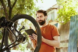 a al aire libre activo vibrante caucásico hombre explorador el Internet para bicicleta mantenimiento. comprometido sano masculino ciclista utilizando teléfono tableta a reparar bicicleta rueda para verano ocio ciclismo. foto