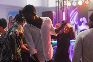 African american clubber in sunglasses partying on crowded dancefloor at nightclub discotheque. Diverse people dancing and moving to electronic music beats at club disco party photo