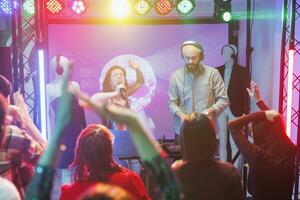 Woman singer engaging crowd to dance and performing on stage with spotlights in nightclub. People enjoying nightlife entertainment and partying on dancefloor at concert in club photo