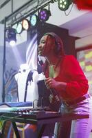 Woman dj interacting with audience and speaking from stage while performing in nightclub. African american musician singing and playing electronic music at discotheque party in club photo