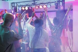Energetic crowd dancing and enjoying discotheque on dancefloor with colorful spotlights. People jumping, having fun and moving to music beats with raised hands in nightclub photo