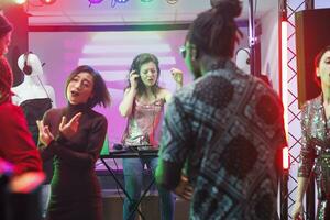 Woman dj in headphones performing using electronic music mixing station on stage while crowd dancing in nightclub. People partying on dancefloor and clubbing at discotheque photo