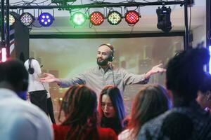 Man dj in headphones performing on nightclub stage at electronic music festival while people dancing. Young musician mixing tracks while clubbers crowd having fun on dancefloor photo