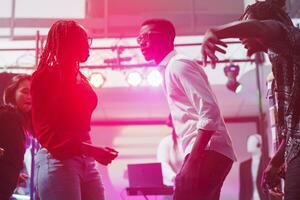 Couple showing improvised dance performance in nightclub illuminated with vibrant lights. Young african american man and woman partying together on dancefloor in dark club photo