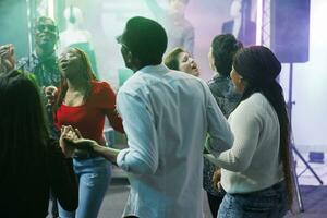 Diverse people clubbing and dancing together at discotheque party event in nightclub. Young friends holding hands, singing and relaxing on crowded dancefloor at social gathering photo