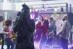 African american man covering ear while trying to chat on mobile phone in nightclub. Young clubber standing on dancefloor and talking on smartphone while music playing loudly in club photo