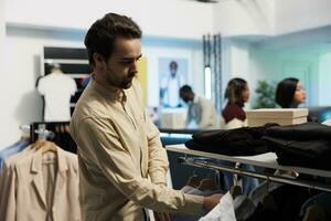 Customer browsing rack with apparel while selecting trendy casual outfit in clothing store. Man exploring hanger with garment while shopping in retail department boutique photo