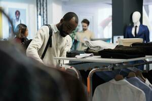 African american man browsing rack with casual clothes while shopping in mall fashion department. Boutique store customer examining menswear apparel while choosing trendy outfit photo