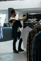 Clothing store customer selecting tie and shirt while shopping for formal outfit. Young man exploring fashion boutique while holding garment and accessory, checking style photo