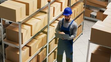 Male supervisor working with laptop in storage room and counting number of boxes on racks. Young adult with hardhat doing stock logistics to ship products orders, merchandise. Handheld shot. photo