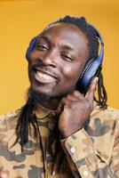 alegre hombre escuchando canción audio, vistiendo auriculares mientras disfrutando música en estudio terminado amarillo antecedentes. africano americano adulto teniendo despreocupado ritmo, bailando durante ocio hora foto