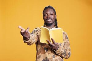 Portrait of cheerful reader being focused on fictional tale during free time, enjoying reading literature book in studio over yellow background. Intellectual man studying for university homework photo
