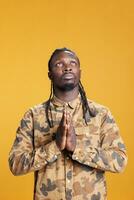 Religious person holding palms together in pray symbol in studio, showing spiritual gesture over yellow background. Faithful african american man praying to jesus for peace and forgiveness. photo