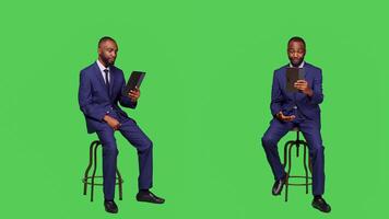Business manager talking on online videoconference call sitting on chair in studio, full body green screen backdrop. Young entrepreneur using tablet with remote videocall meeting. photo