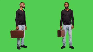 Casual person holding suitcase luggage on camera and waiting for something, standing over green screen backdrop. Young man with briefcase carrying baggage in studio, friendly adult. photo