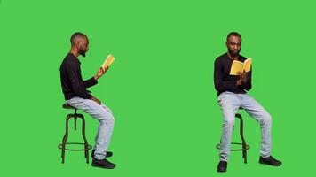 Focused person reading story tale book in studio, sitting on chair over full body greenscreen. Young optimistic man enjoying novel lecture on camera, leisure activity with literature. photo