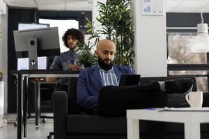 Arabian businessman analyzing financial data on digital tablet in coworking space. Executive manager sitting on couch while working on business development project in corporate office photo